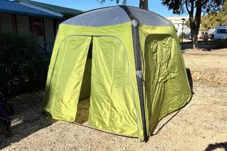 Tenda pop-up verde con la parte anteriore aperta, su ghiaia accanto a un edificio e ad alberi