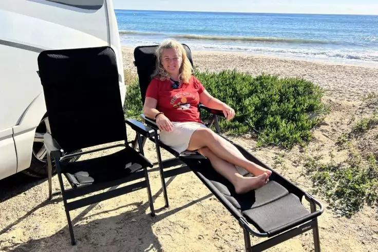 Persona con capelli biondi, indossa una maglia rossa e pantaloncini bianchi, seduta su una sdraio sulla sabbia accanto a un camper, sedia vuota accanto, mare e bassa vegetazione sullo sfondo