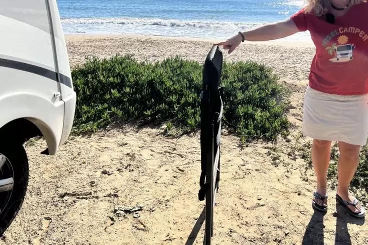 Persona al bordo destro, visibile solo la parte inferiore del corpo, indossa una maglietta rossa, una gonna bianca e sandali, tiene una sedia nera piegata; a sinistra un veicolo bianco, spiaggia sabbiosa con bassa vegetazione e mare sullo sfondo