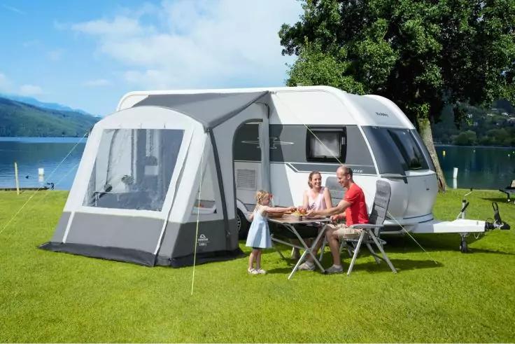 Due adulti e un bambino seduti a un tavolo pieghevole davanti a una roulotte con tenda grigia su un prato vicino a un lago; alberi e montagne sullo sfondo