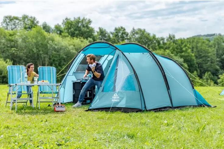 Tenda a tunnel blu su un prato, due persone a un tavolo pieghevole e all'ingresso della tenda, piccolo barbecue/fornello portatile a terra, alberi e cielo sullo sfondo