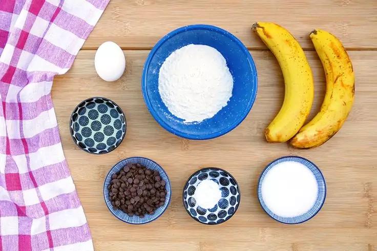 Superficie in legno con strofinaccio a quadri rosa e bianco a sinistra, uovo bianco, grande ciotola blu con polvere bianca, due banane gialle con macchie marroni, quattro piccole ciotole con motivo blu e bianco &mdash; una con pezzetti marroni, due con sostanze bianche, una vuota