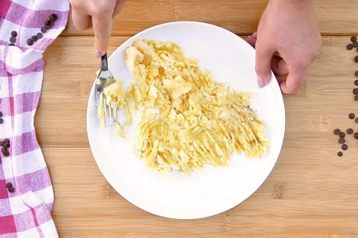 Piatto rotondo bianco con pezzi di frutta gialla schiacciati; mano sinistra regge la forchetta, mano destra sostiene il piatto; panno a quadretti viola a sinistra, gocce di cioccolato a destra su superficie in legno