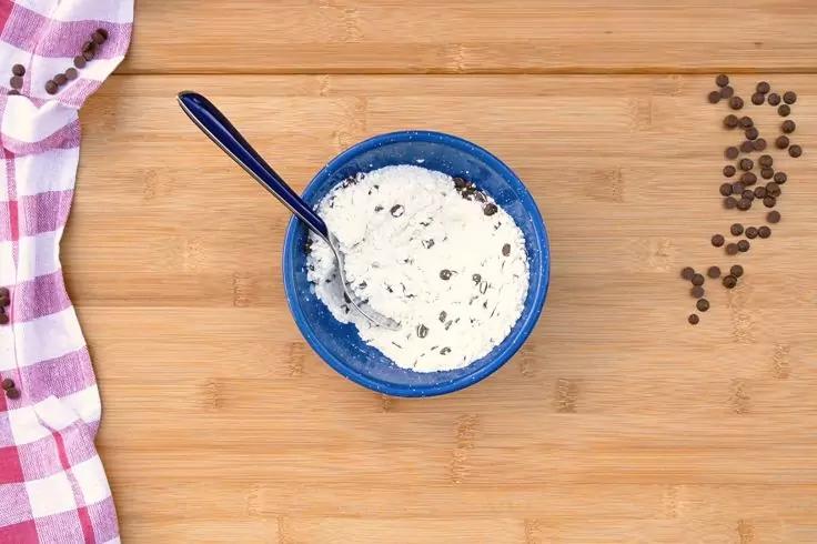 Ciotola blu con massa bianca e cremosa con pezzetti di cioccolato scuro e un cucchiaio su superficie di legno; panno rosa e bianco a sinistra, pezzetti di cioccolato sparsi a destra