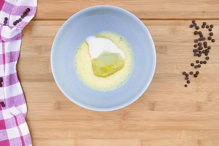 Ciotola azzurro chiaro su tagliere in legno con un pezzo giallo e una sostanza bianca; a sinistra un panno a quadri rosa e bianco, a destra piccoli pezzi rotondi scuri sparsi