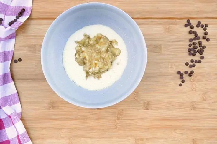 Ciotola azzurro chiaro con liquido bianco e una massa beige e granulosa al centro su una tavola di legno chiaro; a sinistra un panno quadrettato viola e bianco, a destra diversi piccoli pezzi rotondi marrone scuro