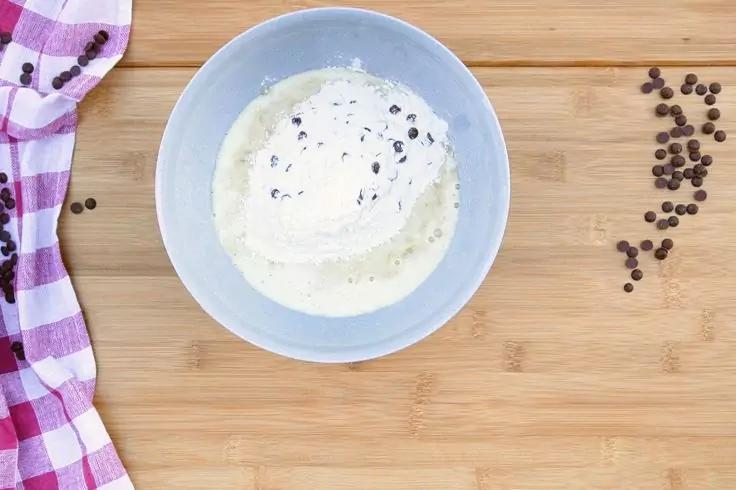 Ciotola azzurro chiaro con una sostanza bianca e cremosa e piccoli pezzi scuri su un tagliere di legno chiaro; a sinistra un panno a quadri viola-bianco, a destra pezzi scuri sparsi