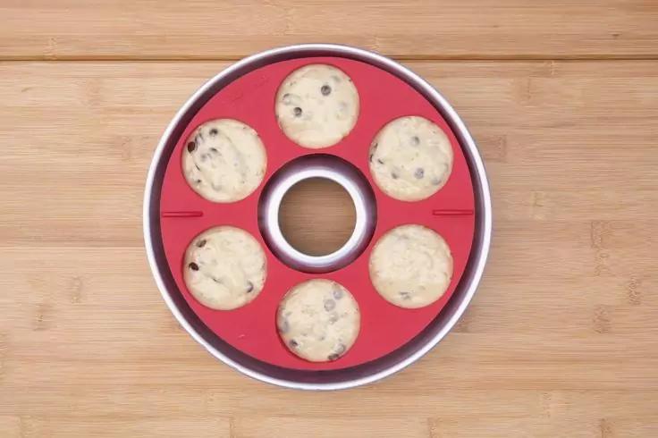 Stampo da forno rotondo rosso con bordo metallico e apertura centrale, riempito con sette porzioni rotonde beige chiaro con pezzi scuri, su una superficie in legno chiaro