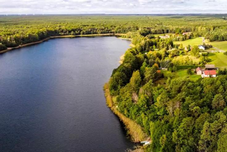Veduta aerea di un lago con riva boschiva, campi e alcune case a destra, cielo nuvoloso