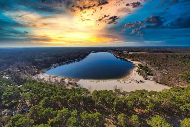 Veduta aerea di un lago con riva sabbiosa, circondato da bosco verde, superficie dell'acqua riflettente, cielo nuvoloso e colorato con il sole vicino all'orizzonte