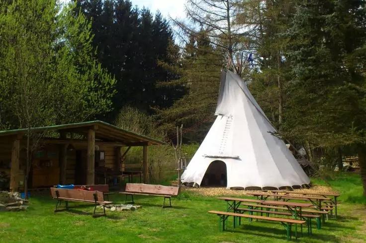 Tipi bianco su una radura erbosa davanti ad alberi sempreverdi, a sinistra un riparo di legno coperto, diverse panchine e tavoli da picnic sull'erba