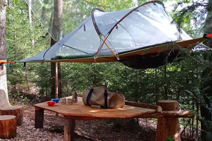 Tenda triangolare trasparente sospesa tra gli alberi sopra un tavolo rotondo in legno; sul tavolo una borsa, due tazze e una borraccia termica; bosco sullo sfondo