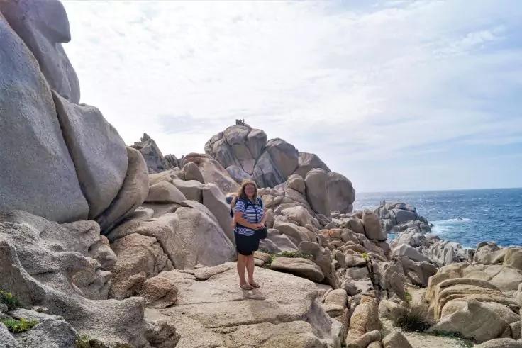 Persona con maglia blu e zaino in piedi su grandi rocce costiere; mare e cielo nuvoloso sullo sfondo