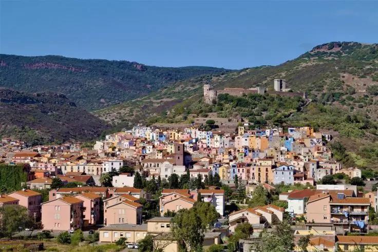 Villaggio collinare con case colorate ravvicinate, una rovina di castello sulla collina e cielo azzurro