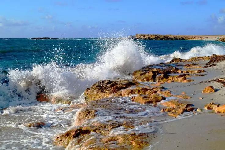 Onde che si infrangono su una costa rocciosa e sabbiosa, spruzzi d'acqua, cielo azzurro