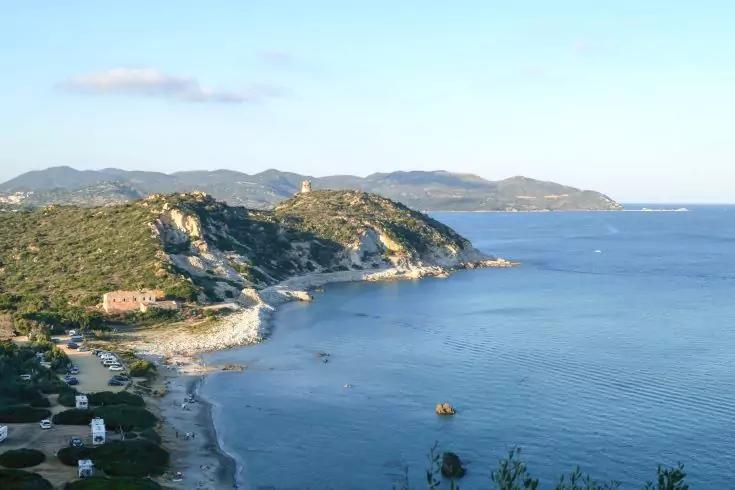 Veduta di una costa con scogliere rocciose, una piccola spiaggia, case sparse, mare calmo e colline sullo sfondo