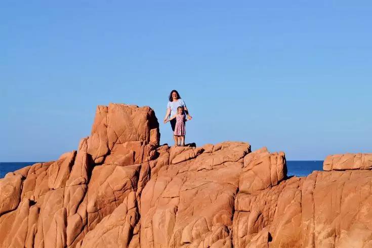 Due persone su rocce rosse vicino al mare sotto un cielo blu sereno; una indossa un top bianco e una gonna chiara, l'altra un vestito rosa