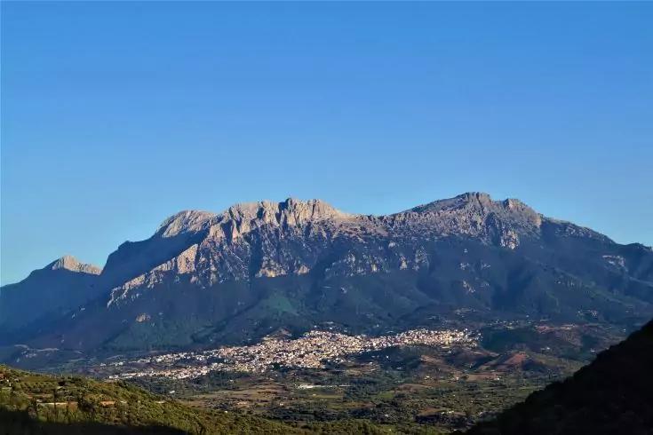 Catena montuosa con cime frastagliate, paese ai piedi delle colline e colline verdi in primo piano sotto un cielo azzurro limpido