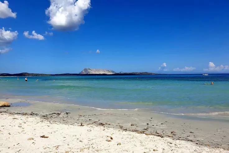 Spiaggia di sabbia con mare turchese poco profondo, piccola isola all'orizzonte, cielo azzurro con poche nuvole e una barca a vela a destra