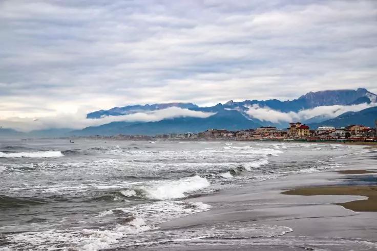 Spiaggia sabbiosa con onde che si infrangono, paese costiero con edifici, montagne sullo sfondo sotto cielo nuvoloso