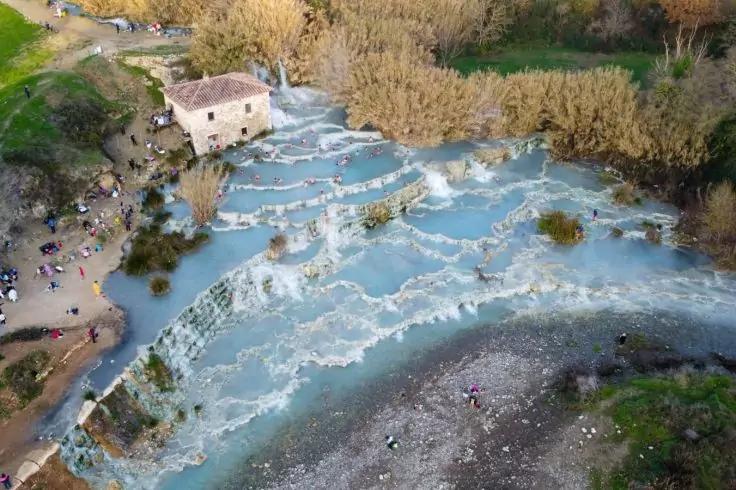 Veduta aerea di vasche terrazzate con acqua turchese lattiginosa in movimento, piccolo edificio in pietra, canneti e persone sulla riva