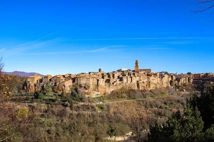 Veduta di un paese su una collina con edifici in pietra ravvicinati e una torre alta, pendii alberati in primo piano, cielo azzurro sereno