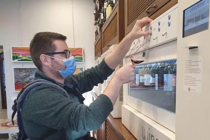 Persona con mascherina e occhiali tiene un bicchiere sotto un erogatore di vino; bottiglie visibili nella macchina, quadri sulla parete