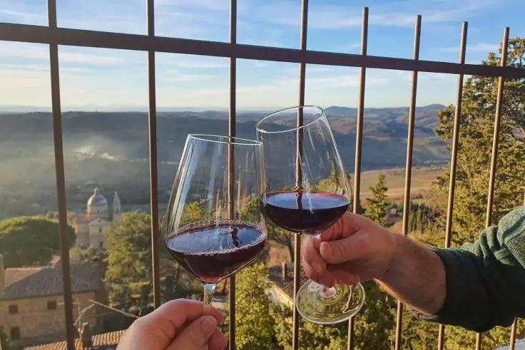 Due coppie di calici di vino rosso, tenuti da due mani, si toccano davanti a una ringhiera di metallo; sullo sfondo nebbia in una valle, colline e un edificio con cupola