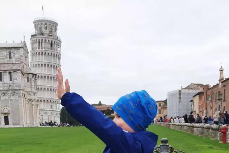 Persona con giacca blu e berretto blu alza un braccio davanti alla Torre di Pisa inclinata; prato e visitatori sullo sfondo