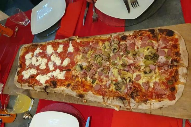 Lunga pizza rettangolare su tagliere in legno, affettata; met&agrave; sinistra con ciuffi di formaggio bianco, met&agrave; destra con sottili fette di carne rosa e funghi affettati; piatti, posate e bevande su una tovaglia rossa visibili