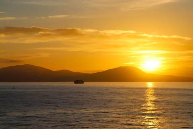 Tramonto su acqua calma con il sole appena sopra le colline, silhouette di una barca e riflesso di luce sull'acqua