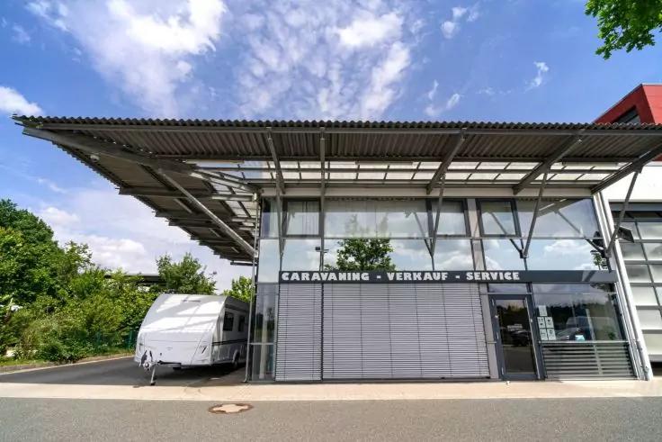 Edificio con facciata in vetro e insegna 'CARAVANING - VERKAUF - SERVICE', ingresso coperto, roulotte a sinistra, strada in primo piano, alberi e cielo nuvoloso