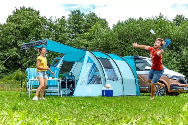 Due persone davanti a una tenda a tunnel azzurro chiaro sull'erba; una persona salta con le braccia alzate, l'altra &egrave; vicino a un tavolo da campeggio. Frigorifero portatile davanti alla tenda, un'auto parcheggiata dietro, alberi sullo sfondo