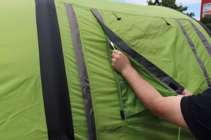 Tenda verde con strisce nere; braccio con mano apre la cerniera di una patta della finestra