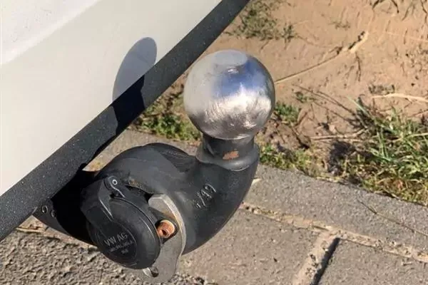 Sfera di gancio di traino argentata su un braccio di gancio nero sotto una parte bianca del veicolo, sopra pavimentazione e terreno sabbioso con erba