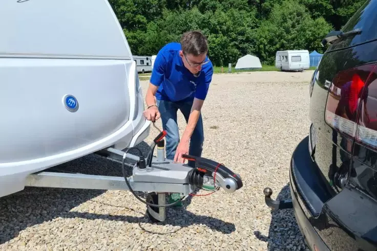 Un uomo con maglia blu collega la barra di traino di una roulotte bianca al gancio di un'auto nera su un piazzale ghiaioso; sullo sfondo altre roulotte e alberi