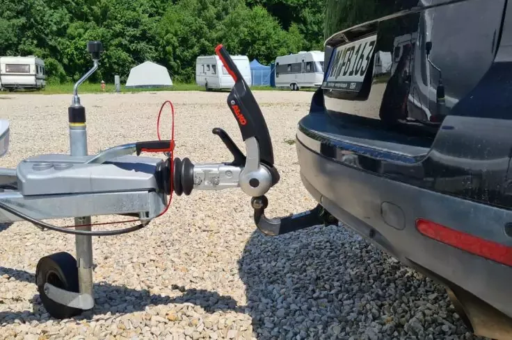Retro di un'auto nera con gancio traino e elemento di accoppiamento montato sulla barra del rimorchio; barra con ruota di supporto, freno a mano e cavo rosso; terreno ghiaioso; roulotte e alberi sullo sfondo
