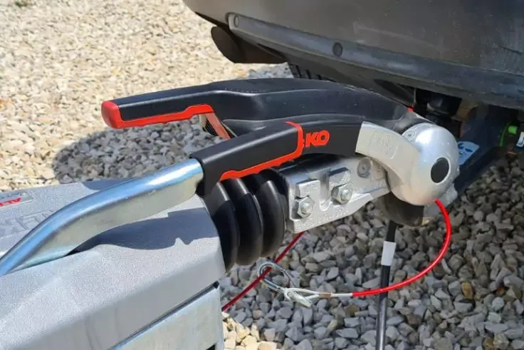 Primo piano di un attacco del rimorchio su una sfera di traino, cavo rosso e connettore, parte del paraurti e terreno ghiaioso visibili