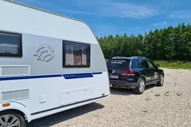 Caravan bianco a sinistra, SUV nero a destra su un'area di ghiaia, alberi e cielo azzurro sullo sfondo