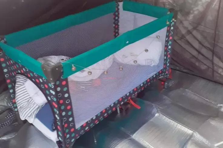 Lettino da viaggio con bordo superiore verde e lati in rete, tessuto a pois colorati, due oggetti di stoffa bianchi all'interno, sottostrato argentato e divano sullo sfondo