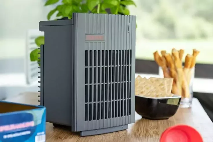 Dispositivo rettangolare grigio con griglie verticali su un tavolo in legno; sullo sfondo una pianta in vaso, una ciotola con cracker e un bicchiere alto con patatine fritte; ciotola rossa e scatola blu sul bordo del tavolo