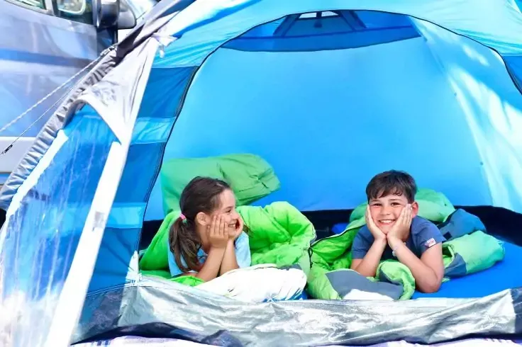 Due bambini sdraiati su materassini all'interno di una tenda da campeggio blu, appoggiando il mento sulle mani e sorridendo
