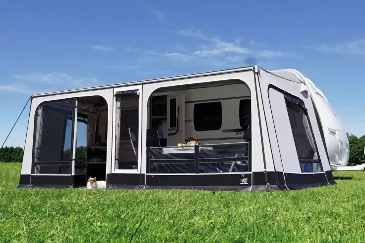 Grande tendalino grigio chiaro fissato a una roulotte bianca su prato verde, finestre visibili e porta aperta, cielo azzurro