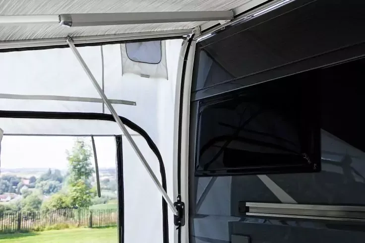 Tetto in tessuto bianco con supporti metallici, televisore nero su una parete scura e, attraverso un pannello laterale aperto, prato, alberi e case sullo sfondo