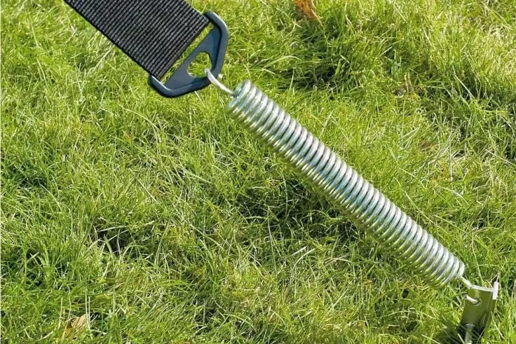 Molla a spirale metallica con ganci, fissata a una cinghia nera e a un picchetto sul prato