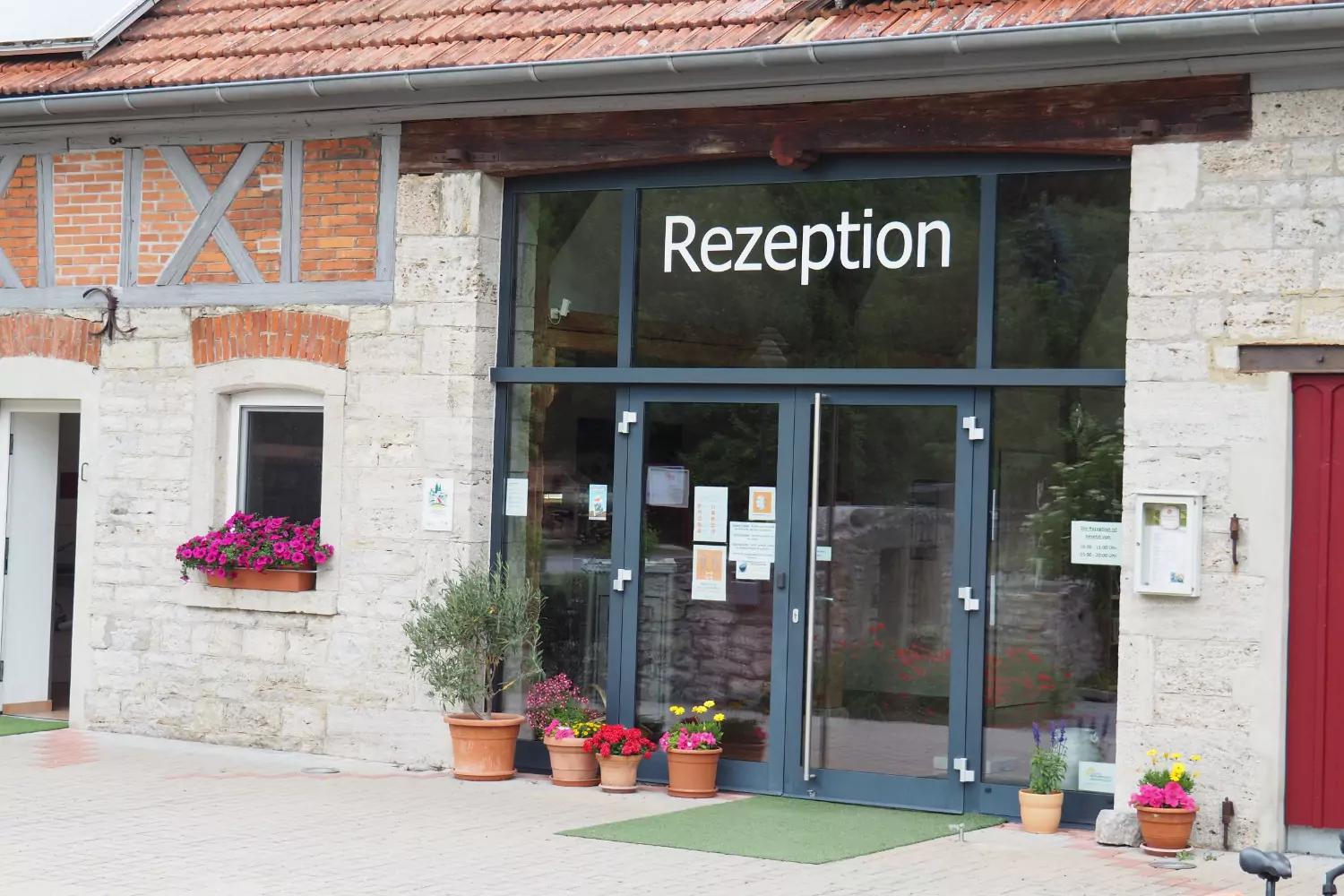 Ingresso con porta in vetro recante la scritta 'Rezeption', facciata in pietra e a graticcio, tetto in tegole, diversi vasi di fiori e una fioriera alla finestra, zerbino verde