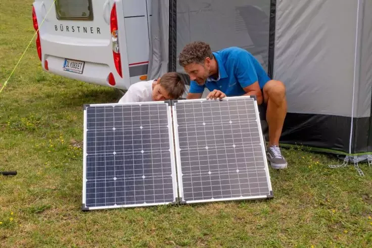 Due persone accovacciate accanto a un pannello solare pieghevole aperto sull'erba; sullo sfondo un camper e una tenda da veranda