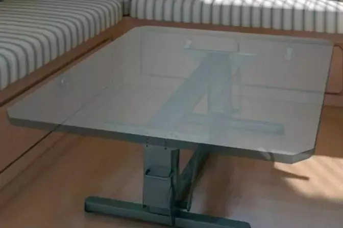 Tavolo quadrato in vetro con base metallica davanti a una panca imbottita con rivestimento chiaro a righe su pavimento in legno, ombra sul pavimento