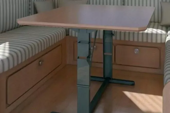 Tavolo rettangolare con piano in legno chiaro e base centrale metallica nera tra due panche imbottite a righe; ante e cassetti in legno sotto le panche, luce solare sul pavimento