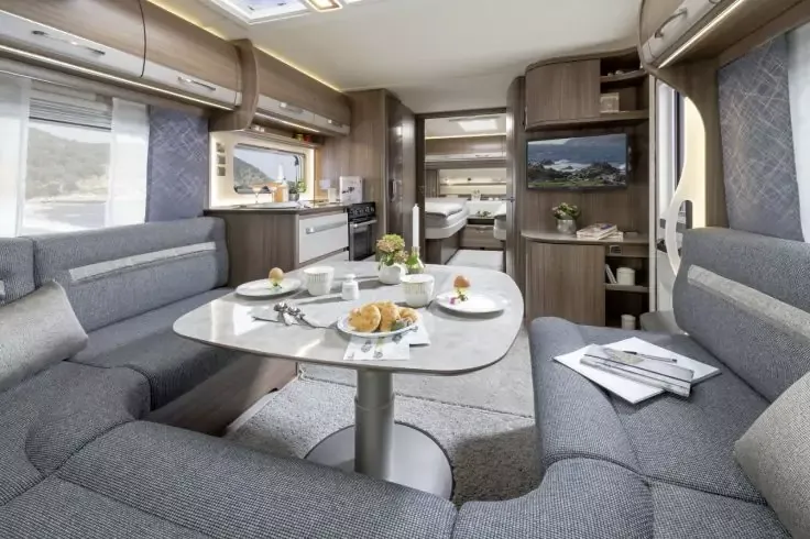 Interno di un camper con divanetto imbottito grigio a U attorno a un tavolo bianco con piatti, tazze, cornetti e un vaso con fiori; angolo cottura con pensili e lavello, televisore a parete e letto sul retro